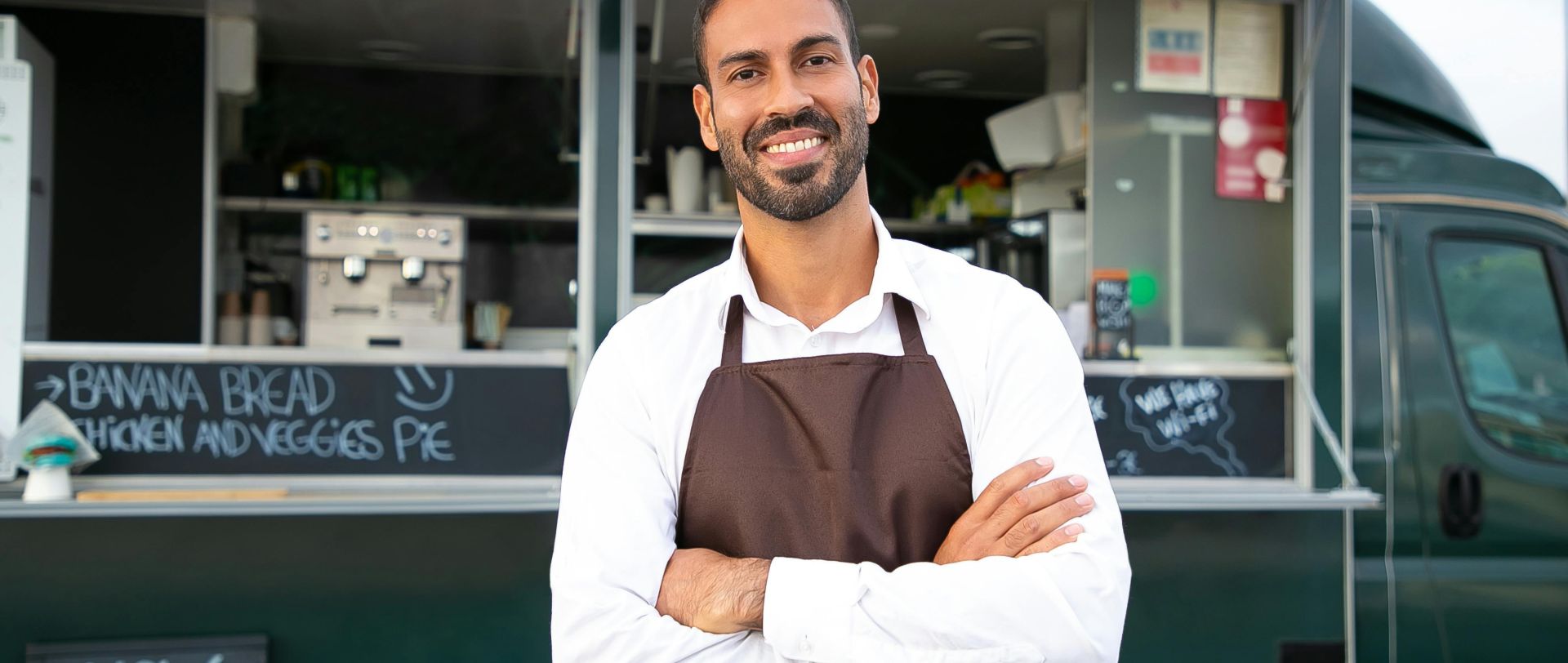 Smiling young ethnic male waiter in apron standing with arms folded near modern food truck and looking at camera contentedly
