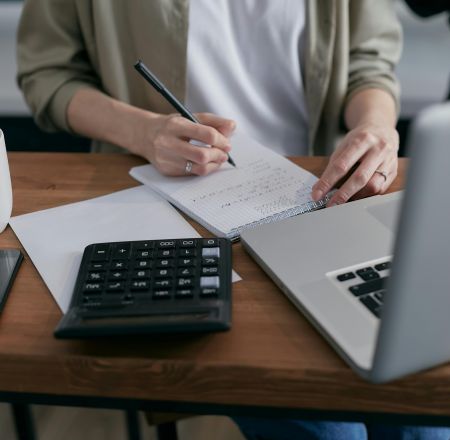 A woman writes financial calculations in a notebook, using a calculator and laptop at a wooden desk.