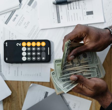Person counting dollar bills over documents with a smartphone calculator on the desk.